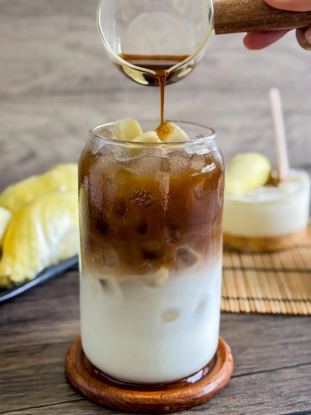Brewed coffee being poured over iced durian coffee topped with durian chunks, showing the layered drink from a side view.