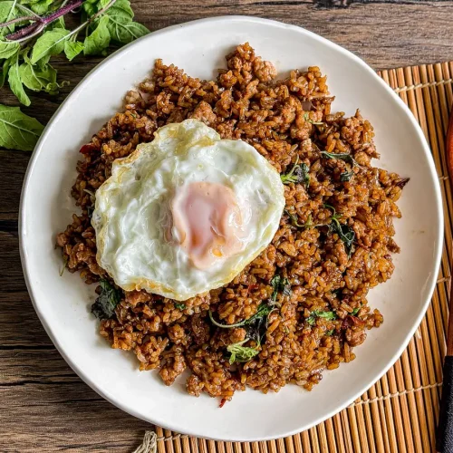 Thai holy basil fried rice with crispy fried egg on top, served on a white dish with a wooden fork and fresh holy basil on the side.