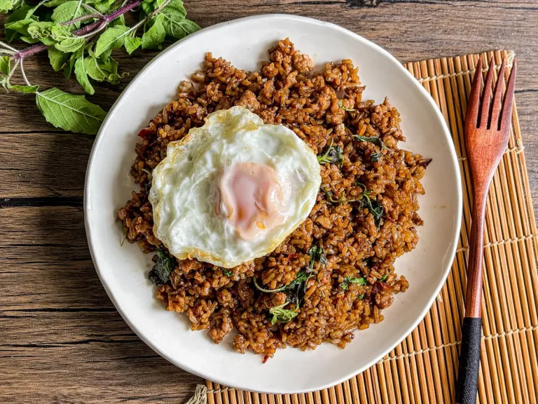 Thai holy basil fried rice with crispy fried egg on top, served on a white dish with a wooden fork and fresh holy basil on the side.