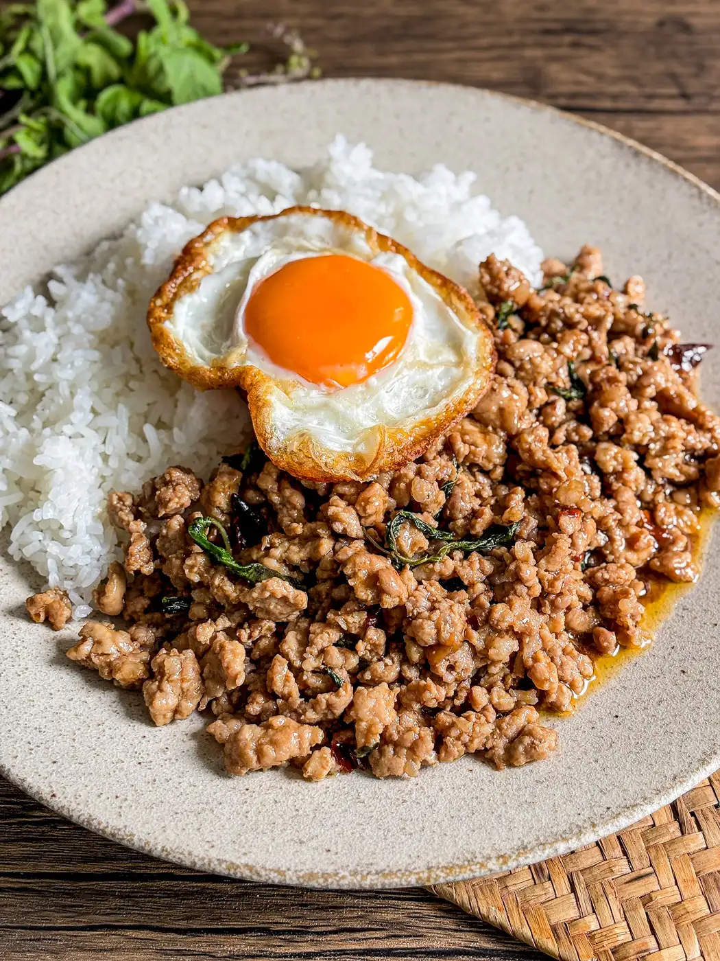 Thai pad krapow with ground pork and holy basil served with jasmine rice and crispy fried egg on a beige plate.