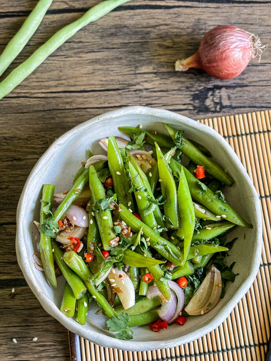 Overhead view of Asian green bean salad with shallots, cilantro, sesame seeds, and sliced red chili in a ceramic bowl.