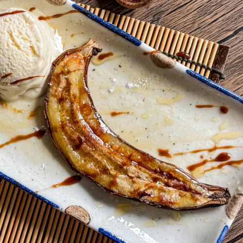 Caramelized air-fried banana served in its peel with a scoop of vanilla ice cream and a drizzle of syrup on a ceramic plate.