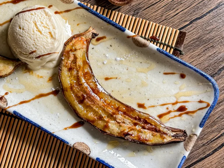 Caramelized air-fried banana served in its peel with a scoop of vanilla ice cream and a drizzle of syrup on a ceramic plate.