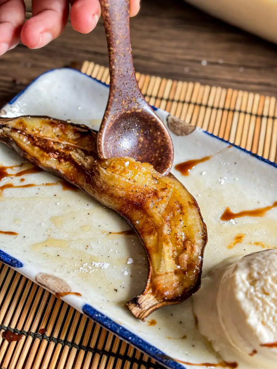 Close-up of a spoon scooping warm, caramelized banana straight from the peel, served with ice cream and syrup.