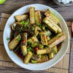 Chinese eggplant stir-fry in a glossy savory sauce, topped with chopped green onions and red chilies, served on a white plate over bamboo mat.