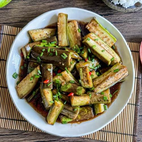 Chinese eggplant stir-fry in a glossy savory sauce, topped with chopped green onions and red chilies, served on a white plate over bamboo mat.