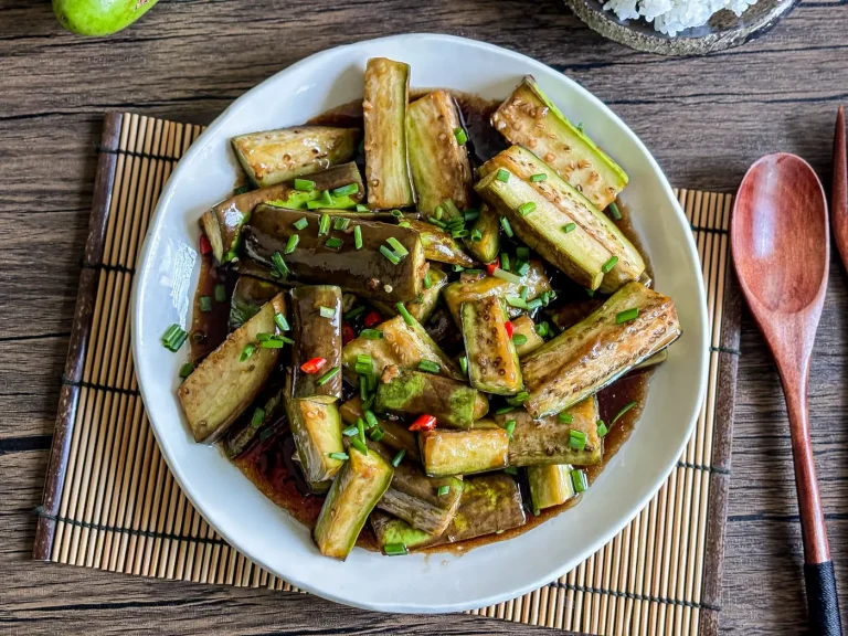 Chinese eggplant stir-fry in a glossy savory sauce, topped with chopped green onions and red chilies, served on a white plate over bamboo mat.