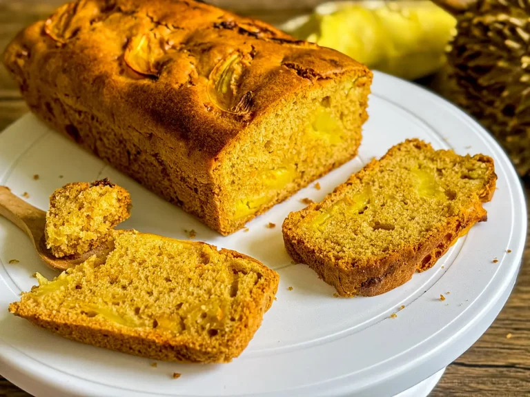 Sliced durian cake loaf on white cake stand with visible durian chunks and a wooden spoon holding a soft bite.
