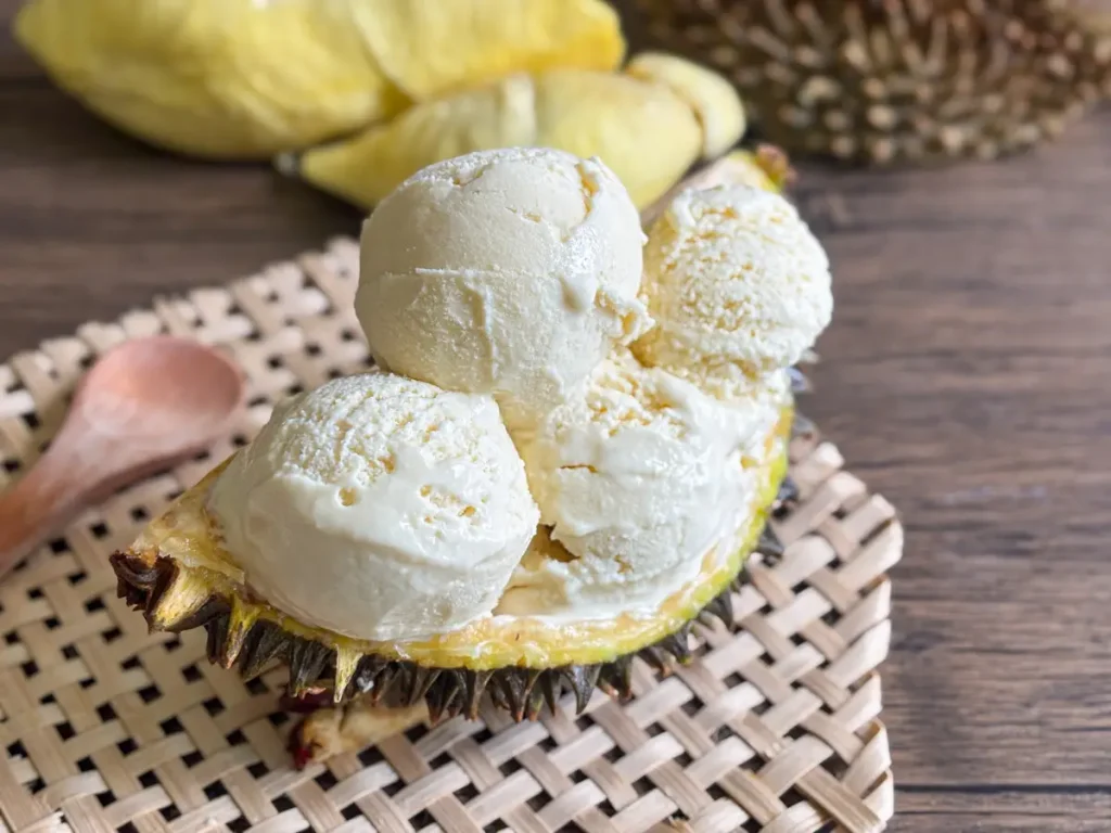 Homemade durian ice cream served in a half durian shell on a woven mat, with wooden spoon on the side.