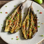 Grilled eggplant halves scored and topped with sesame seeds, sliced red chili, and green onions on a ceramic plate.
