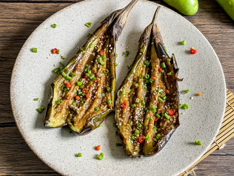 Grilled eggplant halves scored and topped with sesame seeds, sliced red chili, and green onions on a ceramic plate.