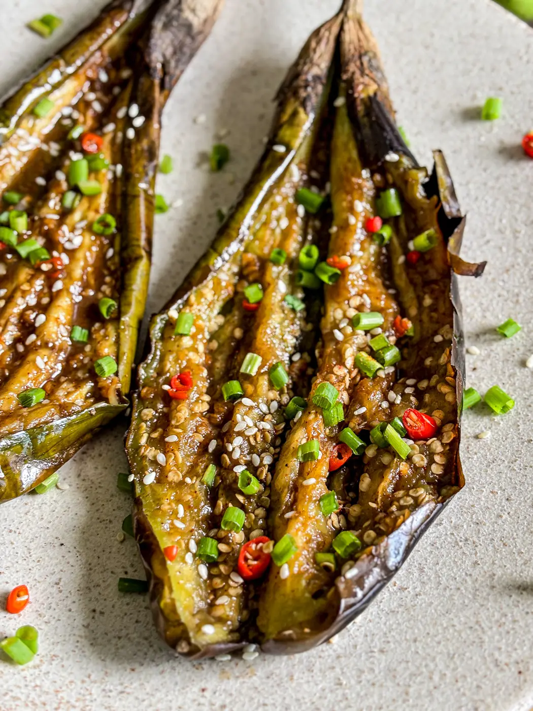 Close-up of grilled eggplant halves garnished with sesame seeds, red chili slices, and chopped green onions on a ceramic plate.