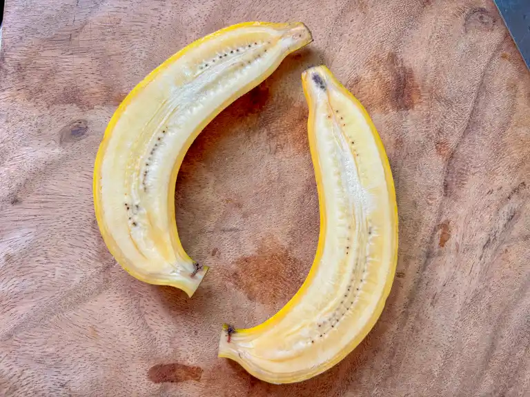 Two raw banana halves placed on a wooden cutting board.