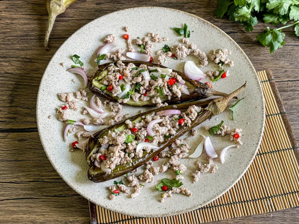 Thai grilled eggplant salad on a ceramic plate with minced pork, red chilies, shallots, and herbs, served on a bamboo mat.