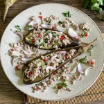 Thai grilled eggplant salad on a ceramic plate with minced pork, red chilies, shallots, and herbs, served on a bamboo mat.