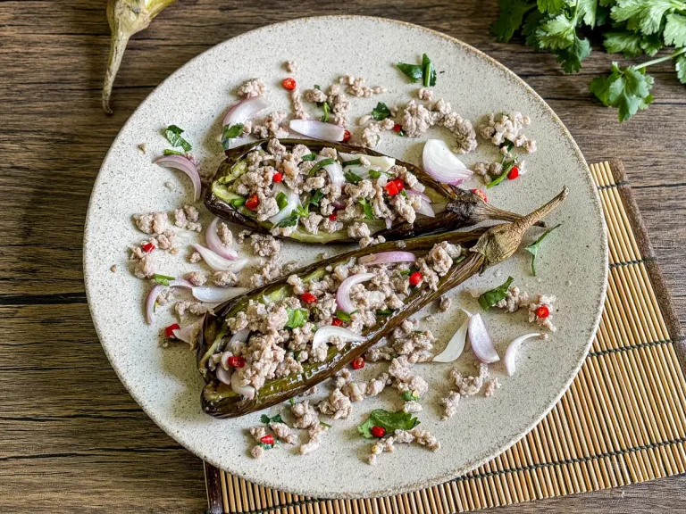 Thai grilled eggplant salad on a ceramic plate with minced pork, red chilies, shallots, and herbs, served on a bamboo mat.