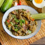Thai roasted eggplant dip with fresh mint, cucumber, and hard-boiled eggs, served on a bamboo mat with vegetables in the background.