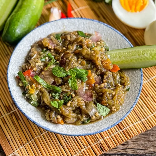 Thai roasted eggplant dip with fresh mint, cucumber, and hard-boiled eggs, served on a bamboo mat with vegetables in the background.