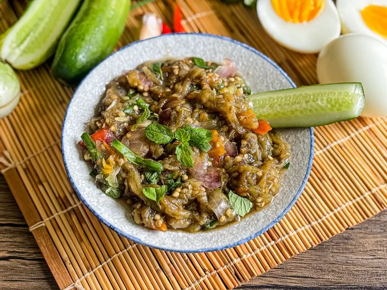 Thai roasted eggplant dip with fresh mint, cucumber, and hard-boiled eggs, served on a bamboo mat with vegetables in the background.