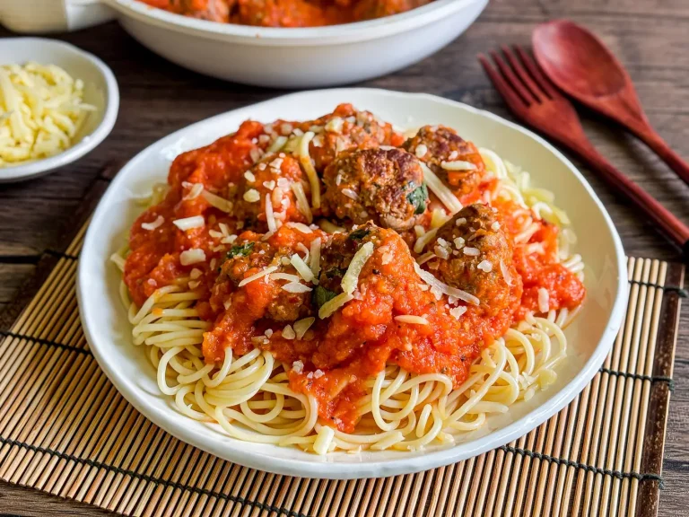 Thai spaghetti meatballs with rich red tomato sauce and shredded cheese served on a white plate over a bamboo placemat with wooden cutlery in the background.