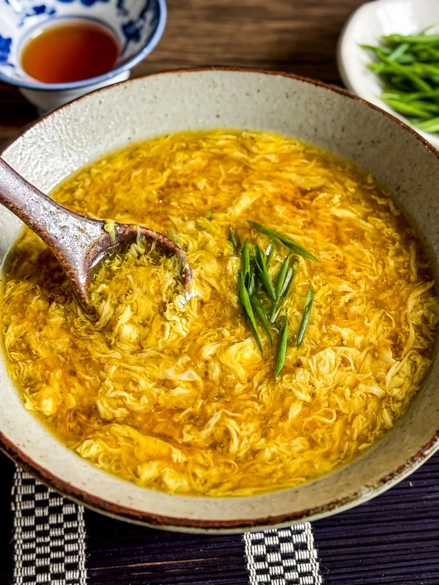 Close-up of egg drop soup in a rustic bowl with a wooden spoon lifting silky egg ribbons, topped with fresh green onions.