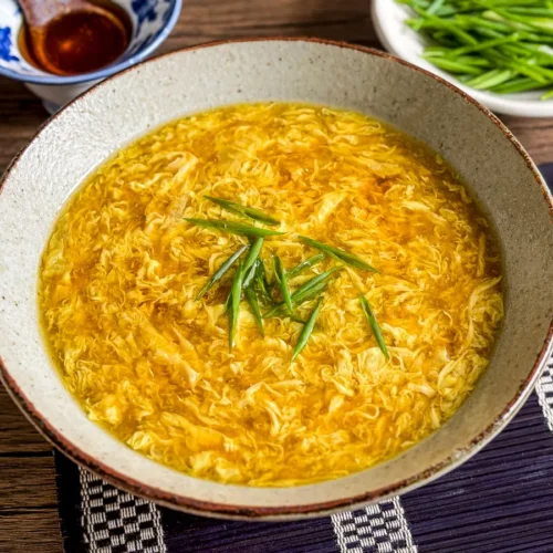 Bowl of egg drop soup topped with sliced green onions, with sesame sauce and fresh chives in the background.