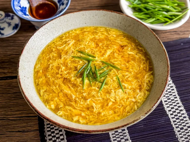 Bowl of egg drop soup topped with sliced green onions, with sesame sauce and fresh chives in the background.