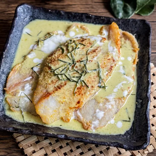Green Thai fish curry with coconut milk and crispy pan-fried fish fillet topped with kaffir lime leaves, served in a square ceramic dish.