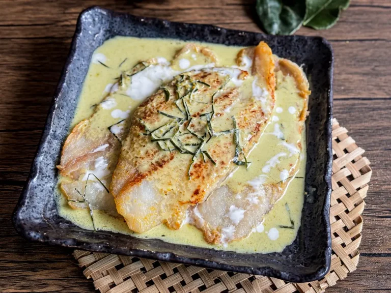 Green Thai fish curry with coconut milk and crispy pan-fried fish fillet topped with kaffir lime leaves, served in a square ceramic dish.