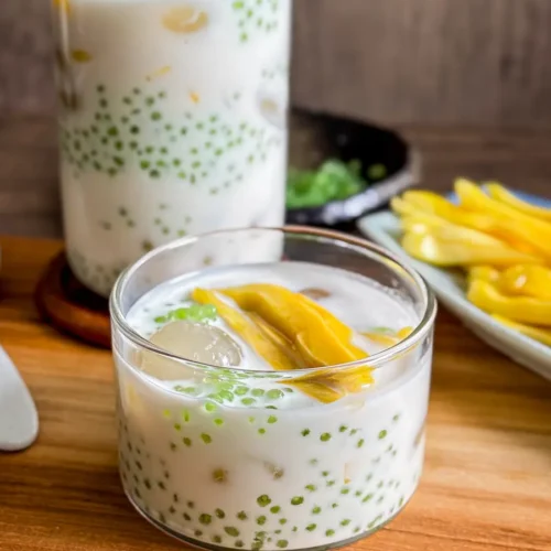 Lao nam wan dessert served in a glass and jar, filled with coconut milk, green sago pearls, longan, and sliced jackfruit, set on a wooden table.