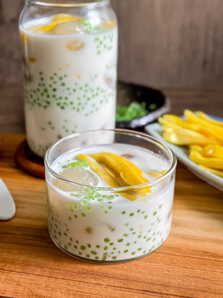 Lao nam wan dessert served in a glass and jar, filled with coconut milk, green sago pearls, longan, and sliced jackfruit, set on a wooden table.