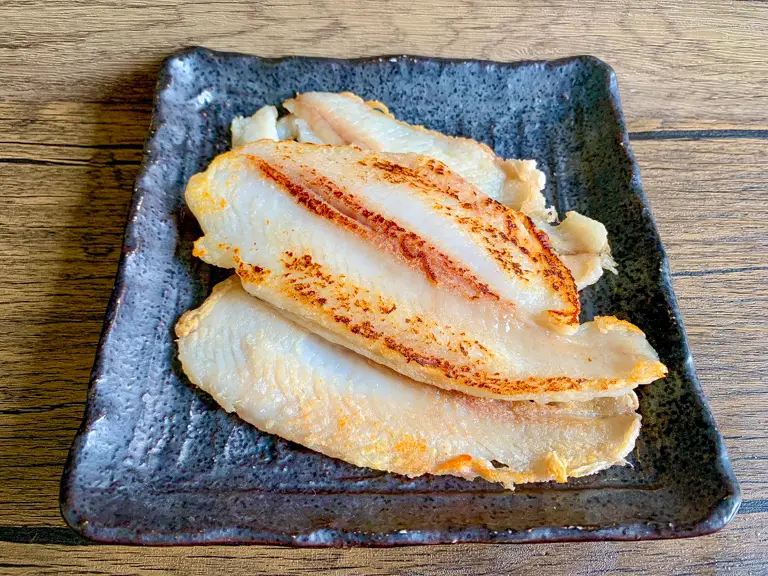 Pan-fried white fish fillets on a dark serving plate, golden and crisp on the edges.