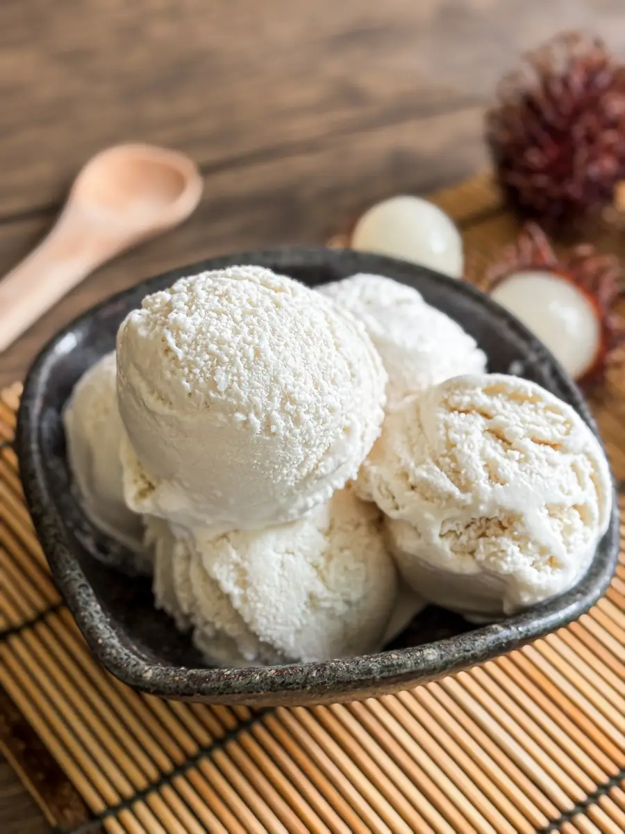 Bowl of homemade rambutan ice cream with smooth, white scoops, placed on a bamboo mat next to peeled rambutans and a wooden spoon.