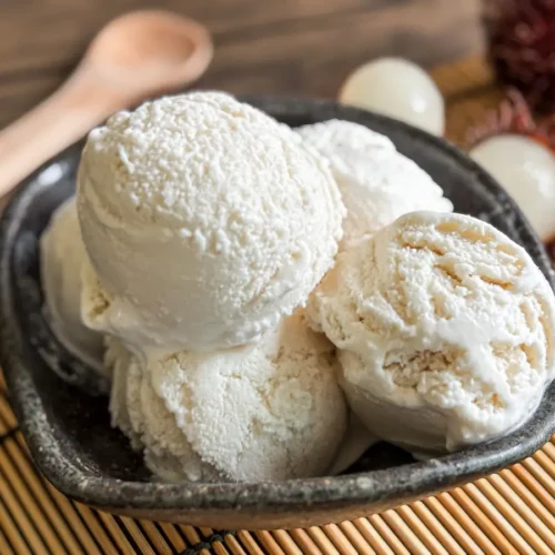 Rambutan ice cream scoops in a dark bowl, served on a bamboo mat with fresh rambutan fruit in the background.