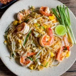 Shrimp pad Thai garnished with garlic chives and a lime wedge, served on a rustic wooden table.