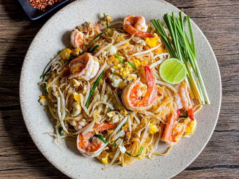 Shrimp pad Thai garnished with garlic chives and a lime wedge, served on a rustic wooden table.