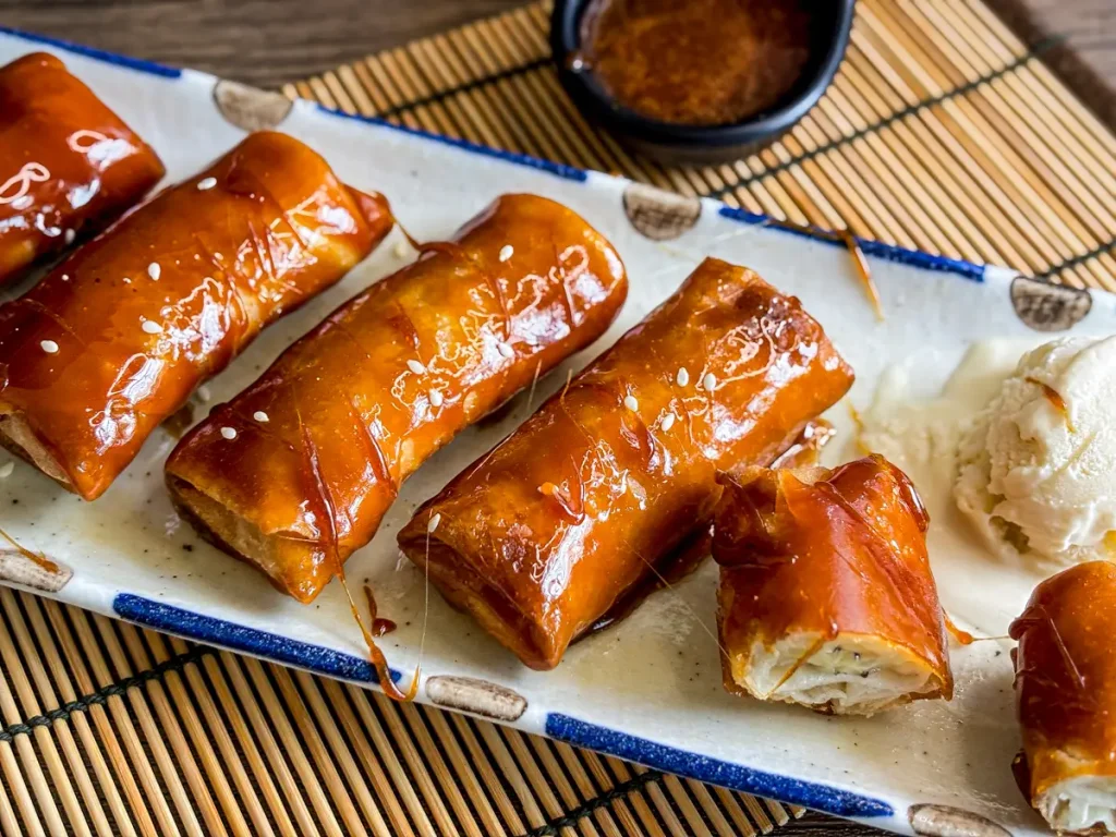 Crispy fried banana lumpia (turon) topped with sticky caramel sauce, served with a scoop of vanilla ice cream on a ceramic plate.