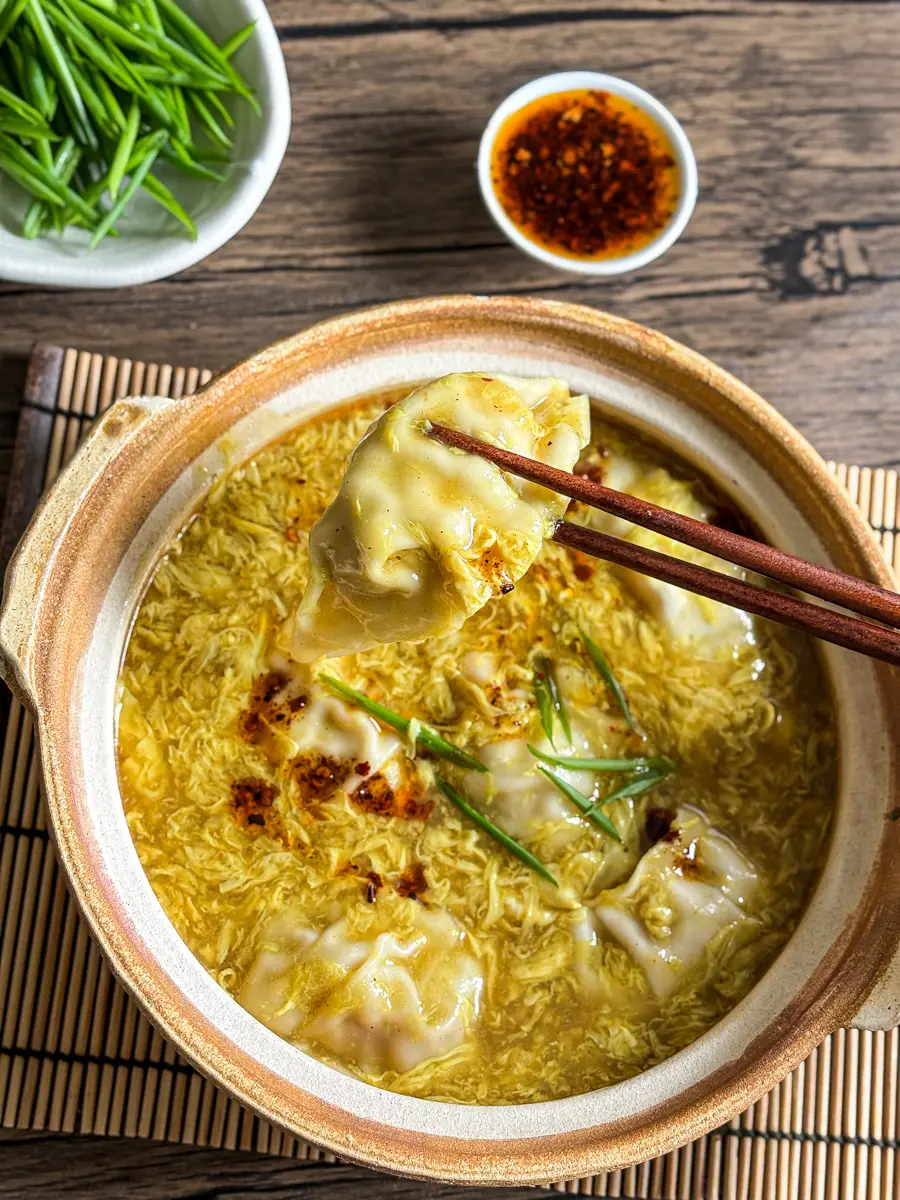 Chopsticks lifting a wonton from a bowl of egg drop soup topped with chili oil and scallions.