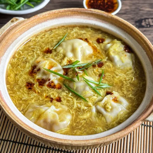 Clay pot filled with wonton egg drop soup, garnished with chili oil and sliced scallions.