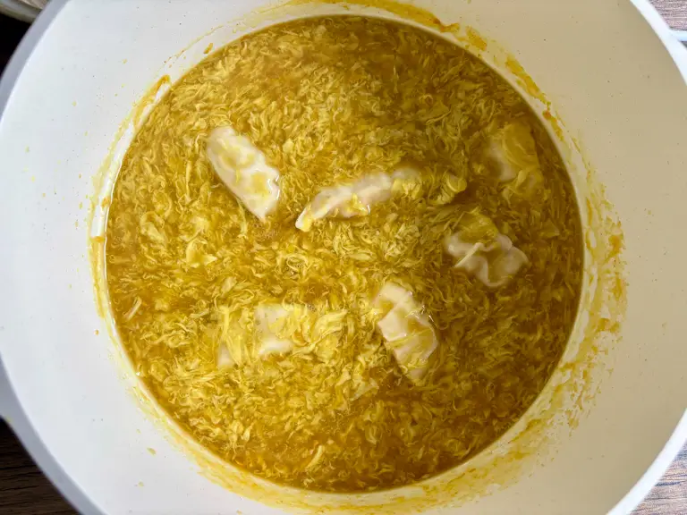 Wontons simmering in golden egg drop soup with silky egg ribbons in a white pot.