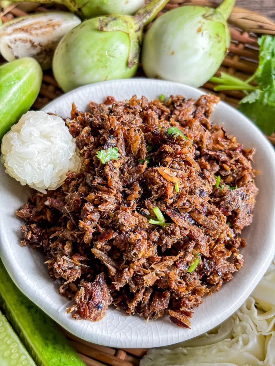 Smoky Lao dried fish dipping sauce (jeow paa) served with sticky rice, cucumber, and Thai eggplants on a woven tray.
