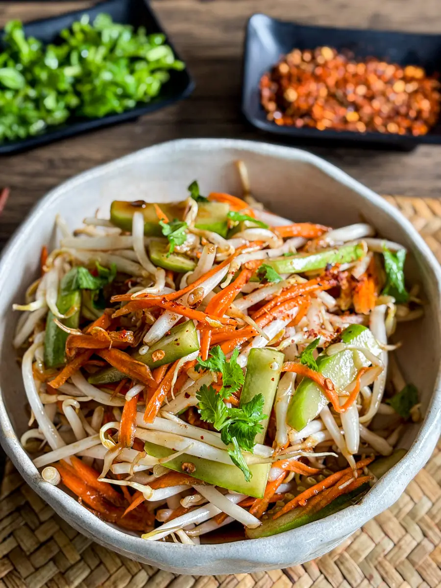 Overhead shot of mung bean sprout salad with fresh vegetables and chili flakes, served cold in a rustic bowl on a woven placemat.