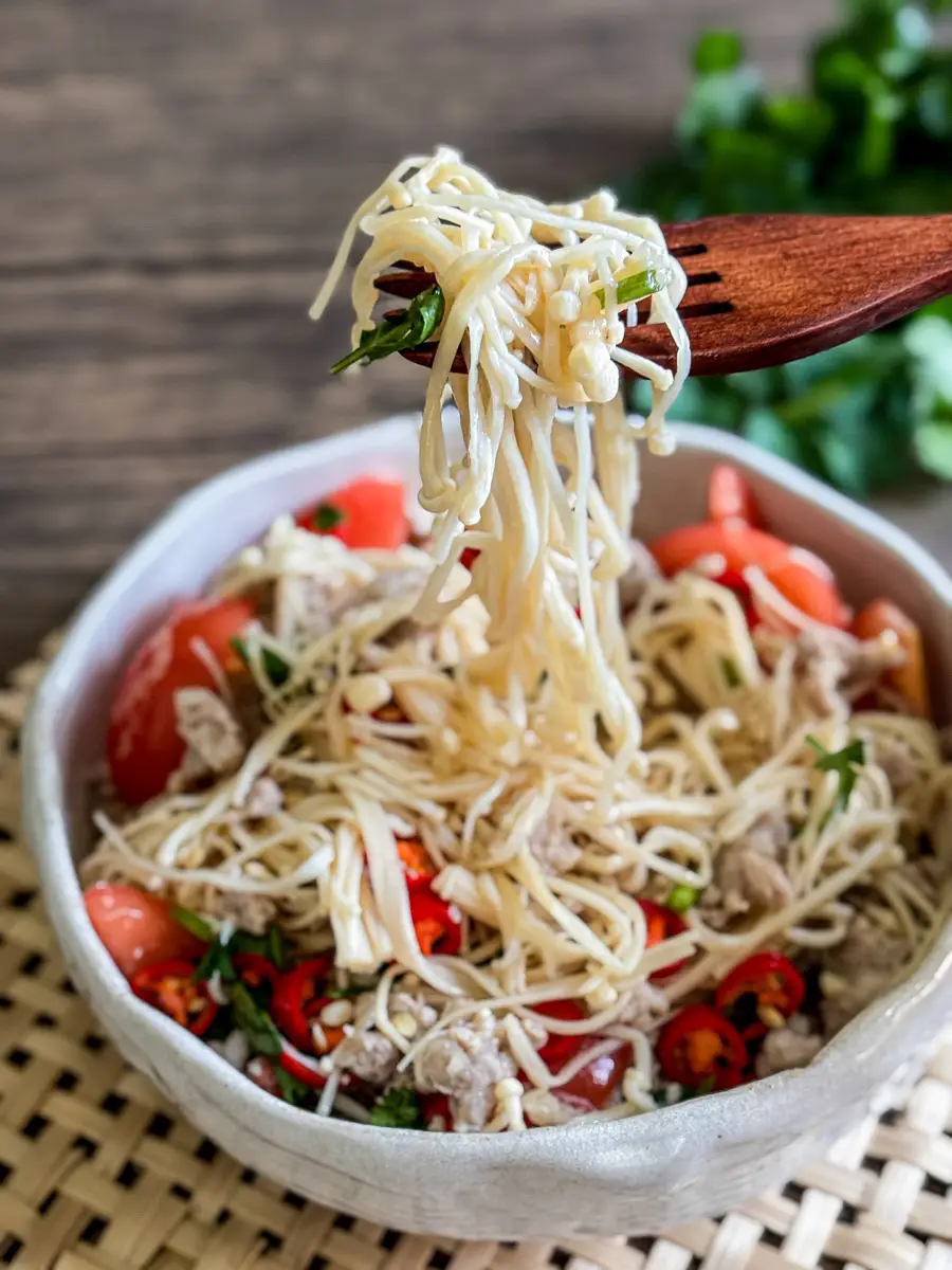 Close-up of wooden fork lifting a bite of cold enoki mushroom salad with coriander and tomato.