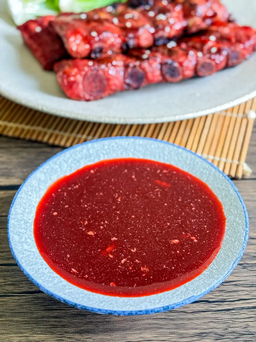Char siu marinade in a blue bowl with red barbecue pork ribs on a plate in the background.
