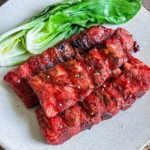 Char siu pork ribs with a sticky, caramelized red glaze, plated with fresh bok choy on a neutral ceramic dish.
