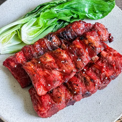 Char siu pork ribs with a sticky, caramelized red glaze, plated with fresh bok choy on a neutral ceramic dish.
