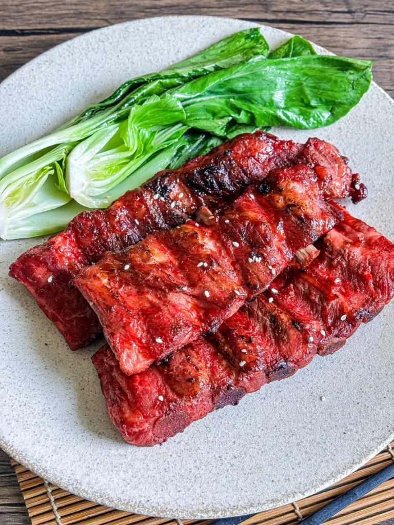 Char siu pork ribs with a sticky, caramelized red glaze, plated with fresh bok choy on a neutral ceramic dish.