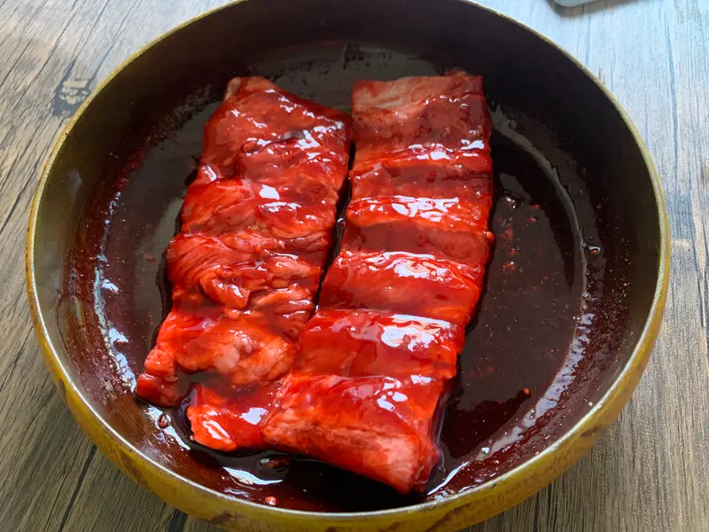 Pork ribs marinating in bright red char siu sauce in a bowl before cooking.