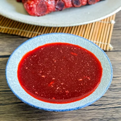 Close-up of homemade red char siu sauce in a small blue bowl on a wooden table.