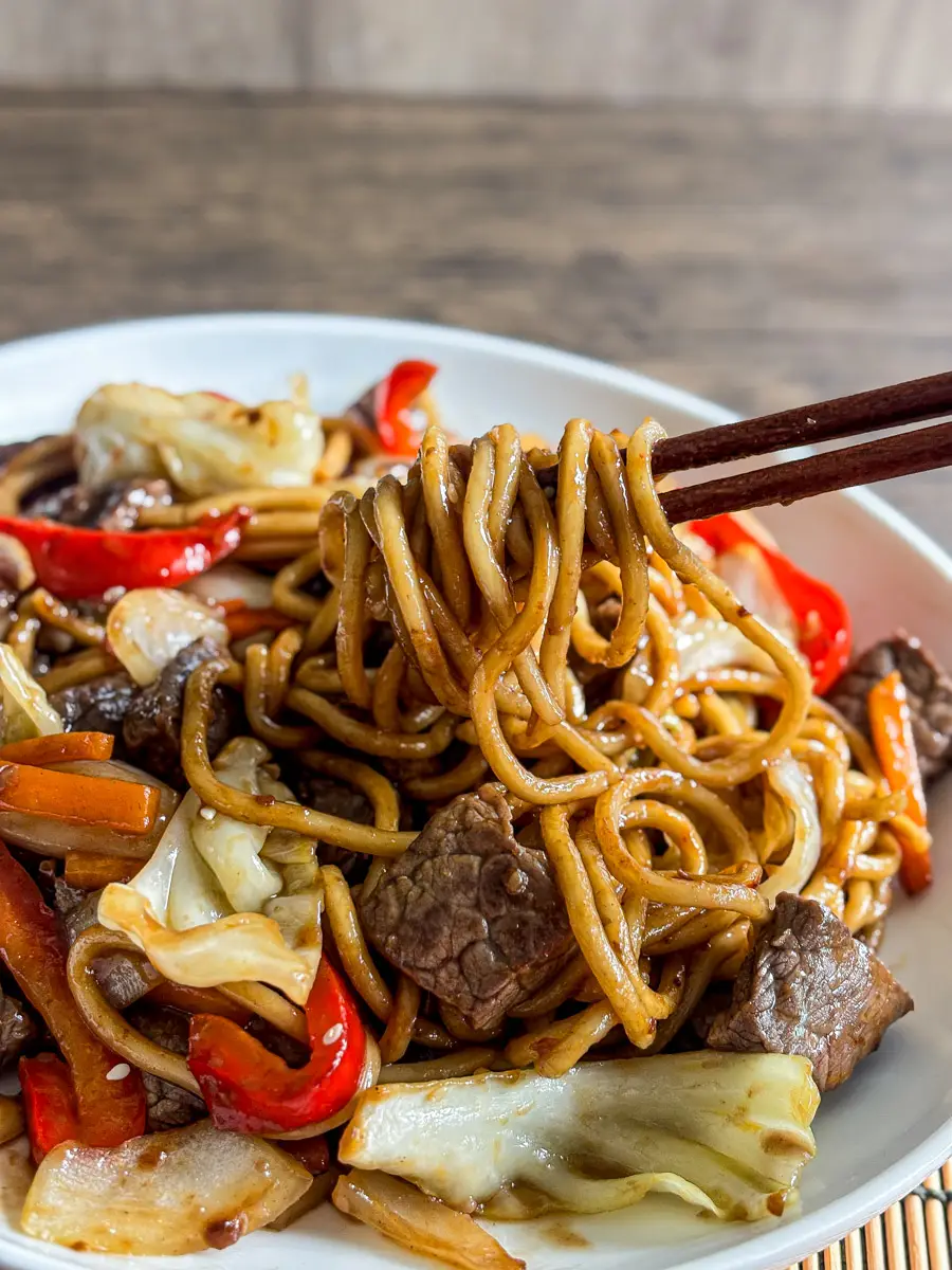 Close-up of beef yakisoba noodles with cabbage, carrots, and bell peppers lifted by chopsticks.